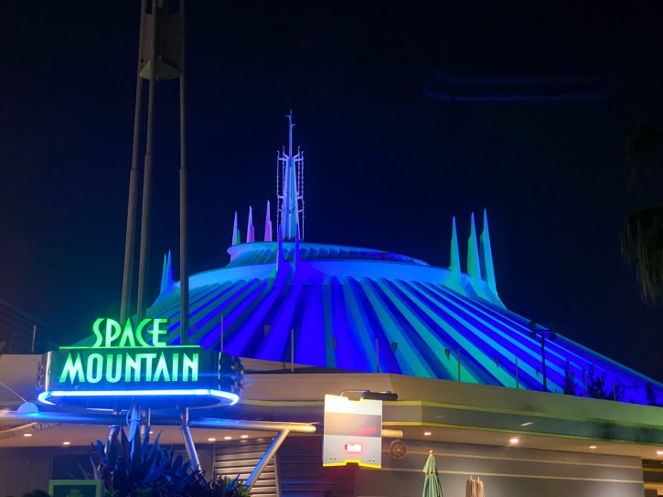 The sign for Space Mountain with the iconic building in the background at night and lit up with blue and green lights.