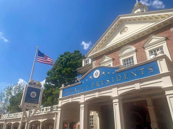 Image from the outside of the Hall of Presidents with a colonial style building and the American Flag. 