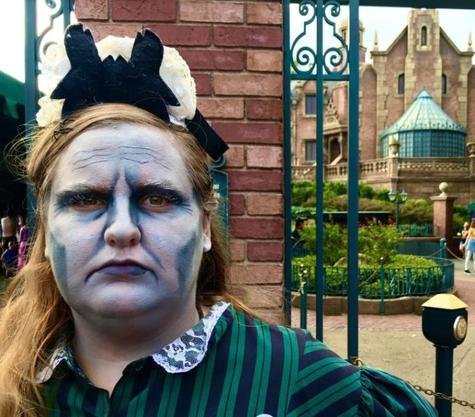 An image of a cast member with ghost makeup on in front of the Haunted Mansion.
