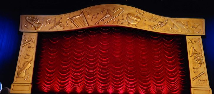The stage at Mickey's PhilharMagic before the show begins. A big red curtain is closed and the area surrounding the curtain is covered in gold reliefs of classical instruments.